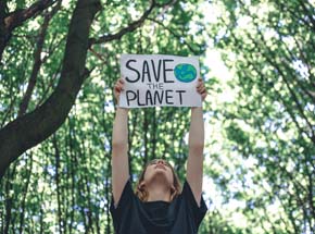 Frau hält Schild mit der Aufschrift "Save the Planet" mit beiden Armen nach oben. Sie steht in einem Wald.