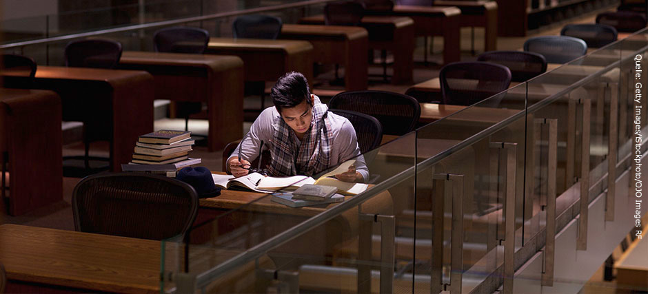 Junger Mann sitzt einsam in einer alten Bibliothek an einem Tisch und arbeitet ein Buch durch. Auf dem Tisch liegt zu seiner rechten Seite ein Stapel Bücher.