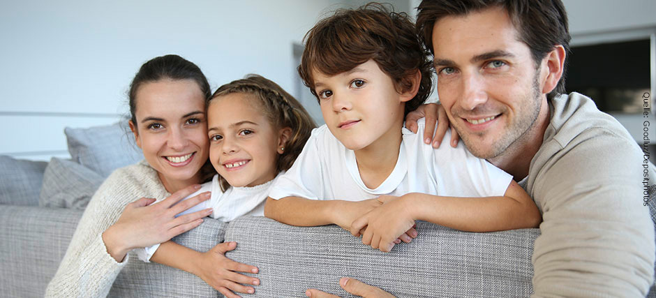 Eine glückliche junge Familie mit Sohn und Tochter sitzt auf einem Sofa und schaut über die Rückenlehne ins Bild.