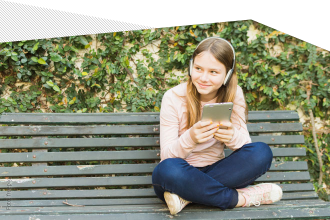 Mädchen sitzt im Schneidersitz auf einer Bank im Park und hält ein Smartphone in der Hand. Auf ihrem Kopf hat sie Kopfhörer. Die